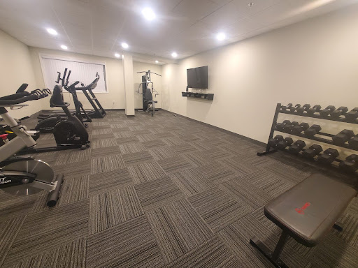 An empty fitness room with boring beige walls and grey flooring.  Cardio machines line the left side of the room, an all-in-one weight machine is at the back next to a window, a large TV on the right hand side with freeweights and a bench in the foreground.